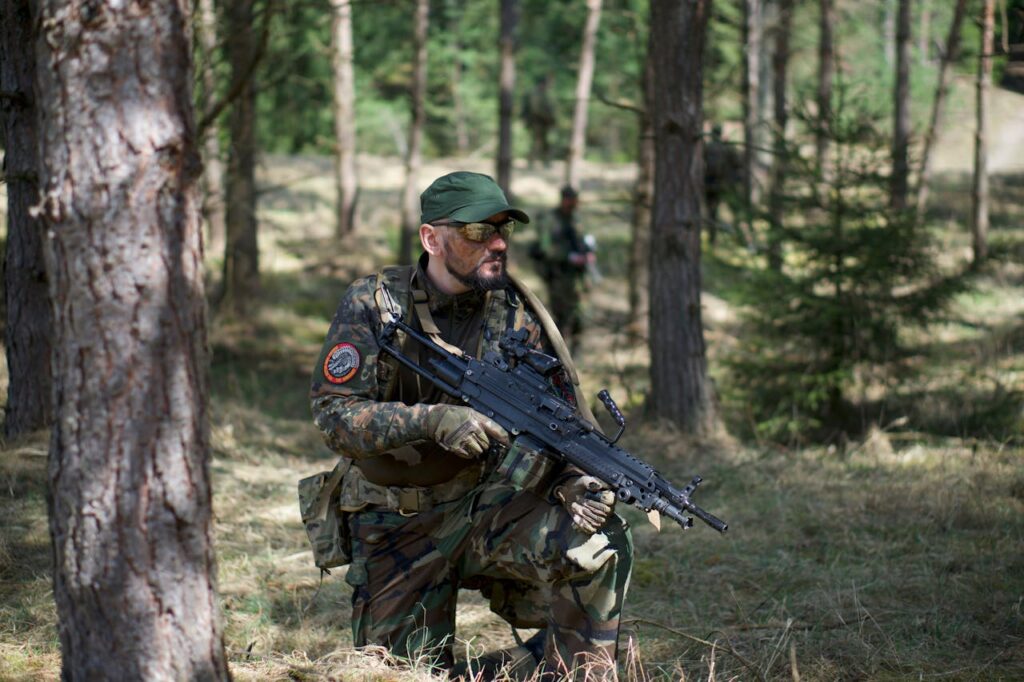 A soldier in camouflage gear crouching in a forest, armed with a rifle, ready for action.